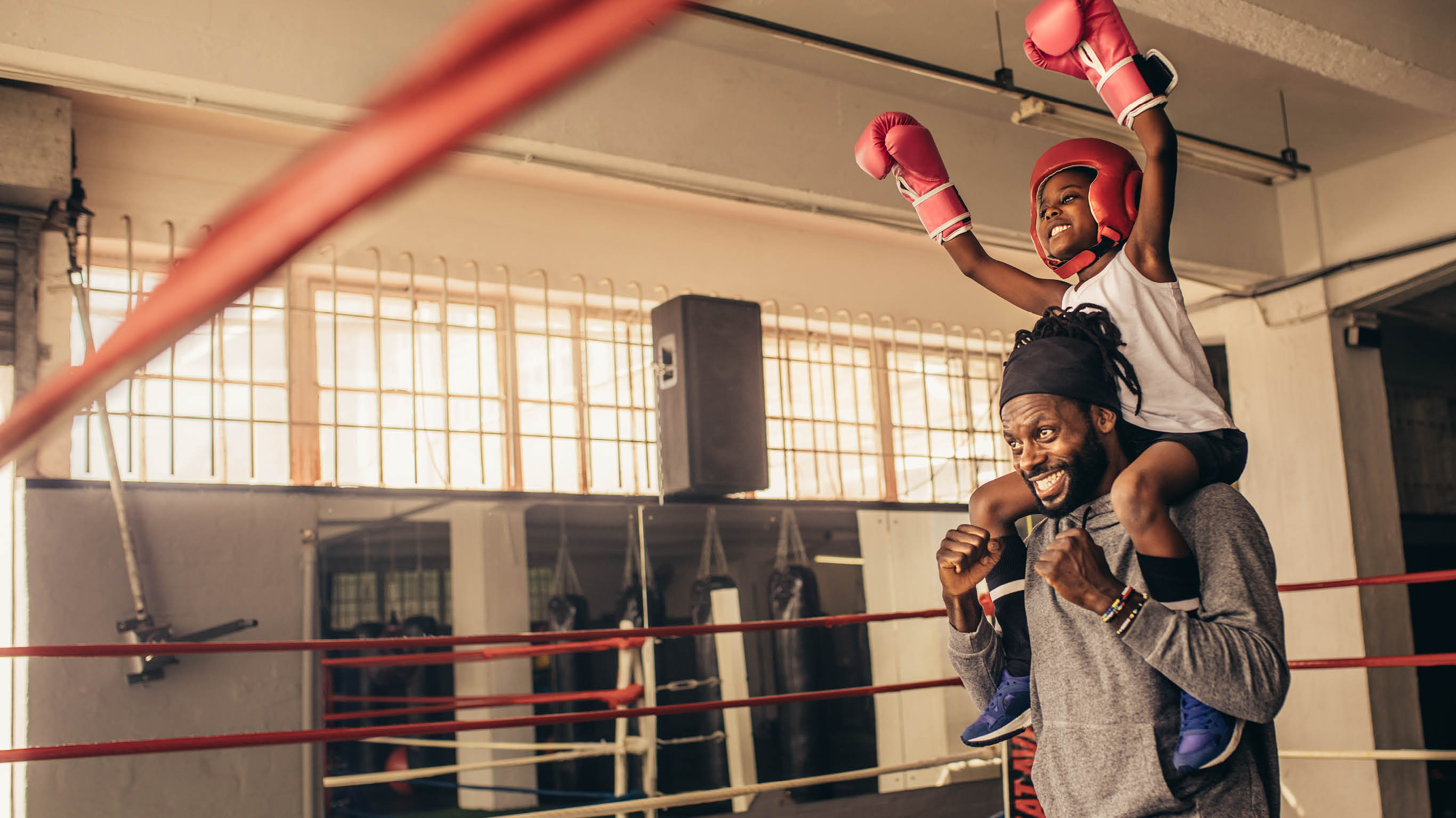 Trainer carrying a boxer kid on shoulders celebrating a win  Kid in boxing gear sitting on shoulder of coach with raised hands 