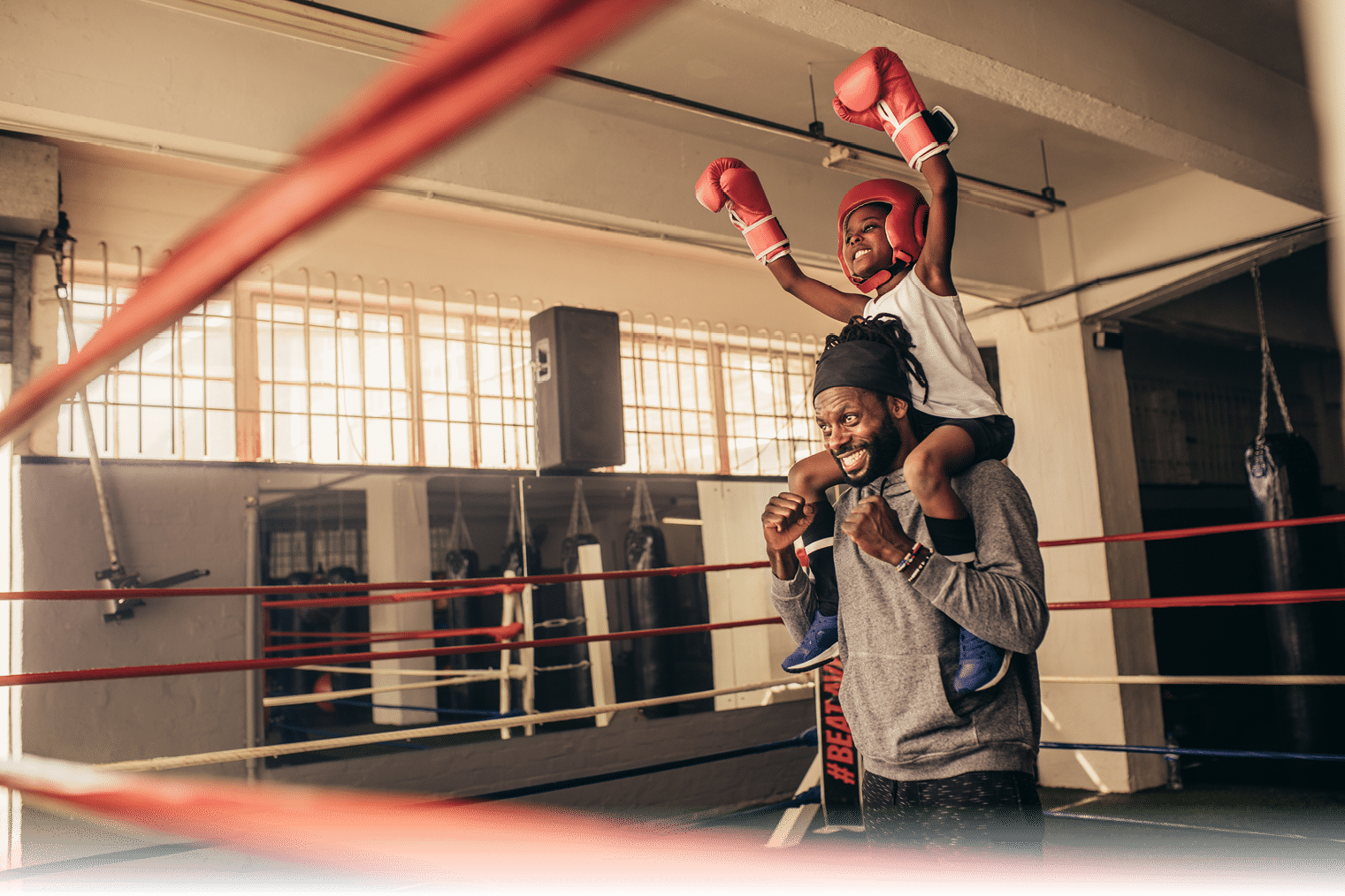 Trainer carrying a boxer kid on shoulders celebrating a win  Kid in boxing gear sitting on shoulder of coach with raised hands 